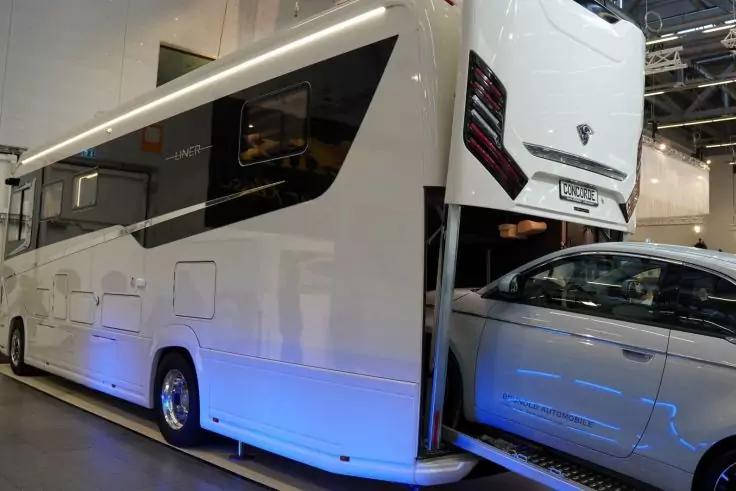 Großes weißes Fahrzeug mit Fenstern und seitlichen Klappen, geöffnetes Heckladefach, darin ein silbernes Auto teilweise geladen; blaue Bodenbeleuchtung; Messehalle im Hintergrund