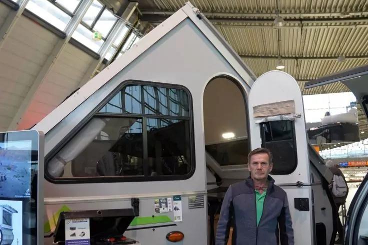 Reisemobilanhänger mit aufgestelltem Dach und geöffneter Tür, ein Mann steht davor, im Hintergrund eine große Halle mit Metallträgerdecke und ein Bildschirm links