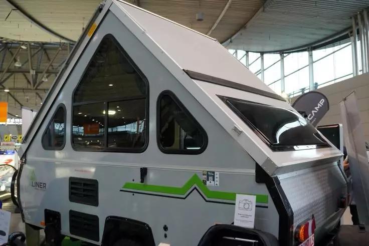 Kleiner grauer A-förmiger Anhänger mit dreieckigen Fenstern, grüner Zierleiste, Lüftungsöffnungen und Rücklicht, in einer Messehalle mit großen Fenstern