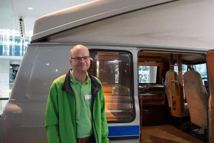 Eine Person mit grüner Jacke und Brille steht neben der geöffneten Tür eines grauen Campingbusses; im Bus sind Sitze sichtbar