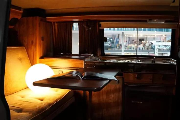 Innenraum eines Wohnmobils mit Holzverkleidung, gepolsterter Sitzbank, runder leuchtender Lampe auf der Bank, Tisch mit offenem Buch, Küchenzeile und Fenster mit Blick auf Straße und Personen