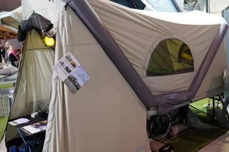 Beige-graues aufgestelltes Zelt mit rundem Fenster, offenem Eingang, Informationsblättern an der Außenseite und Personen im Hintergrund