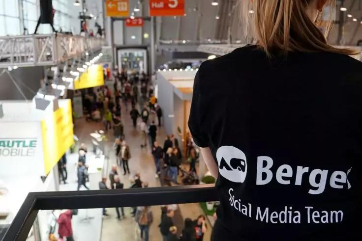 Rückenansicht einer Person mit schwarzem T‑Shirt mit weißem Logo und Schrift 'Berger Social Media Team', an einem Geländer; im Hintergrund eine belebte Messehalle mit Ständen, hängenden Schildern und Personen