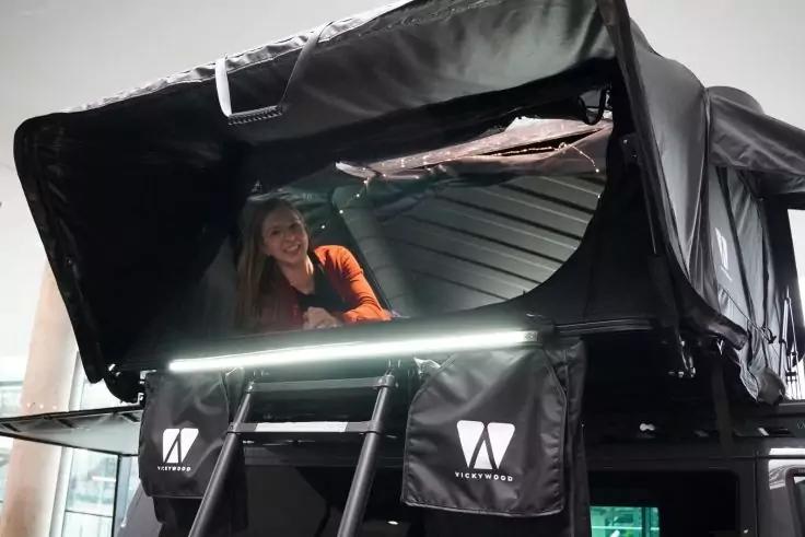 Geöffnetes schwarzes Dachzelt auf einem Fahrzeug mit Leiter, LED-Lichtleiste, zwei Staufächern mit weißen Logos und einer Person in oranger Kleidung im Inneren