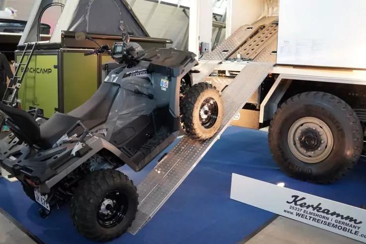 Dunkelgraues ATV auf einer Metallauffahrrampe zum offenen Laderaum eines weißen Lastwagens; sichtbare Räder, blauer Boden und ein Schild am Boden