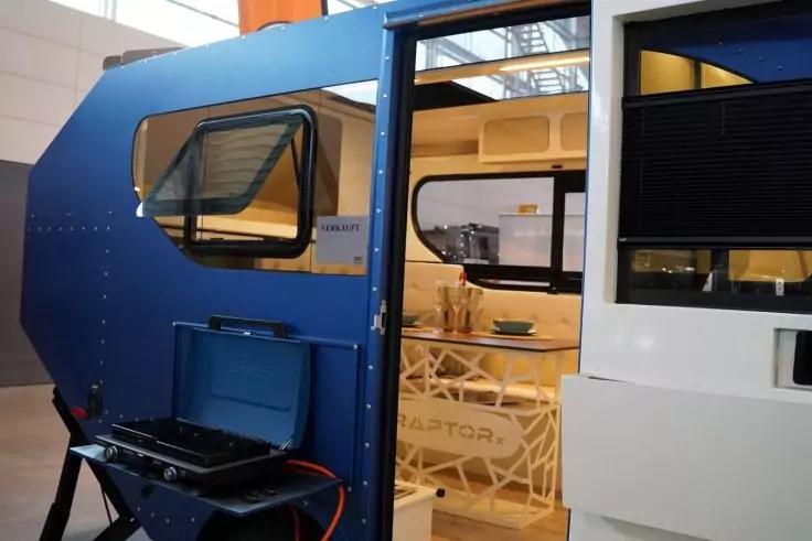 Geöffnete seitliche Tür eines blauen Wohnwagens, Blick ins Innere mit Holztisch, Stühlen und Geschirr; kleiner Grill mit Schlauch außen sichtbar