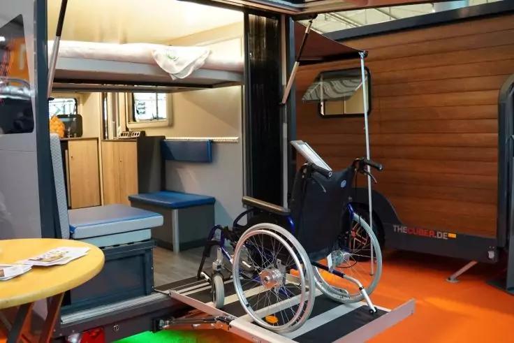 Rollstuhl auf einer Metallrampe vor dem Eingang eines Wohnmobils; innen sichtbar Hochbett, blaue Sitzpolster, Holzwand, gelber Tisch und oranger Boden