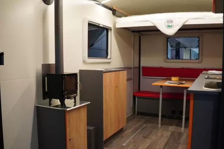 Innenraum mit kleinem Holzofen links, Holzschränken, Fenster, roter Sitzbank mit Tisch und Küchenzeile mit Spüle