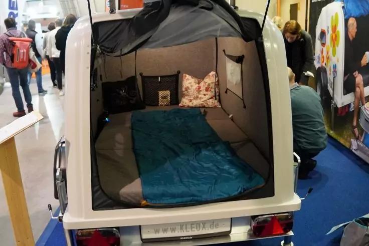 Kleiner weißer Anhänger mit geöffneter Heckklappe, Innenraum mit Matratze, blauem Schlafsack und gemusterten Kissen, Messestand und mehrere Personen im Hintergrund