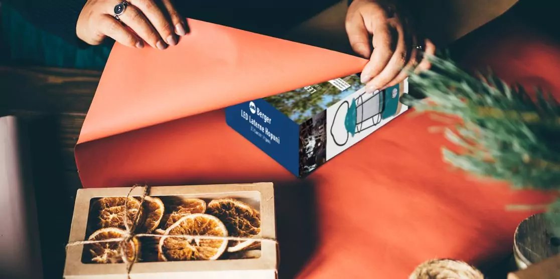 H&auml;nde &ouml;ffnen orangefarbenes Papier &uuml;ber einer Schachtel mit getrockneten Orangenscheiben auf einem roten Tisch, daneben eine blaue Schachtel und ein gr&uuml;ner Zweig