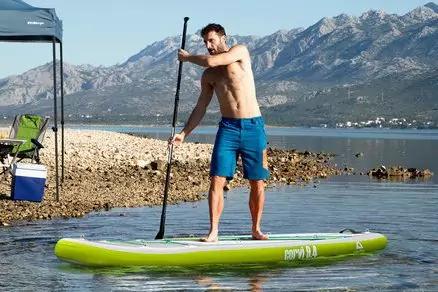 Ein Mann in blauen Shorts steht auf einem grünen Stand-up-Paddleboard und hält ein Paddel; im Hintergrund felsiger Strand, ein Sonnendach und Berge
