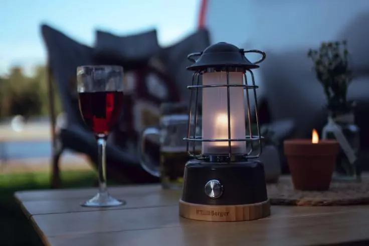 Auf einem Holztisch eine brennende Laterne, daneben ein Glas Rotwein, im Hintergrund unscharfe Stühle, ein kleiner Tontopf mit Pflanze und eine brennende Kerze