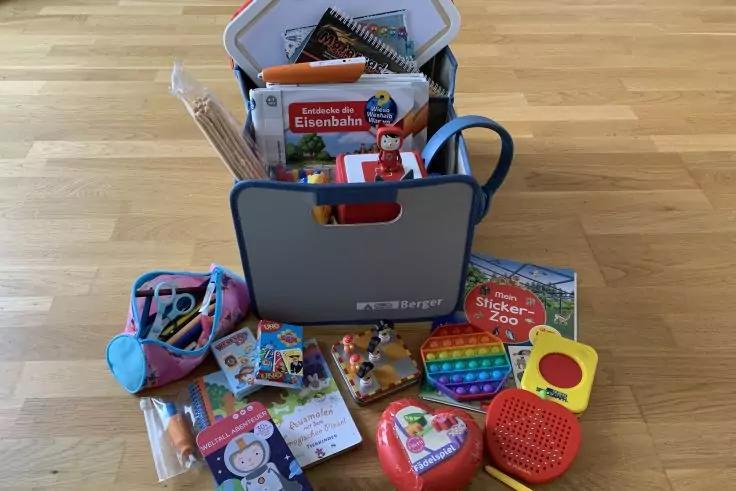 Grauer Aufbewahrungskorb auf Holzfußboden, gefüllt mit Büchern und Spielzeug; davor liegen bunte Spielsachen, ein Regenbogen-Pop‑it, Stifte und eine herzförmige Dose