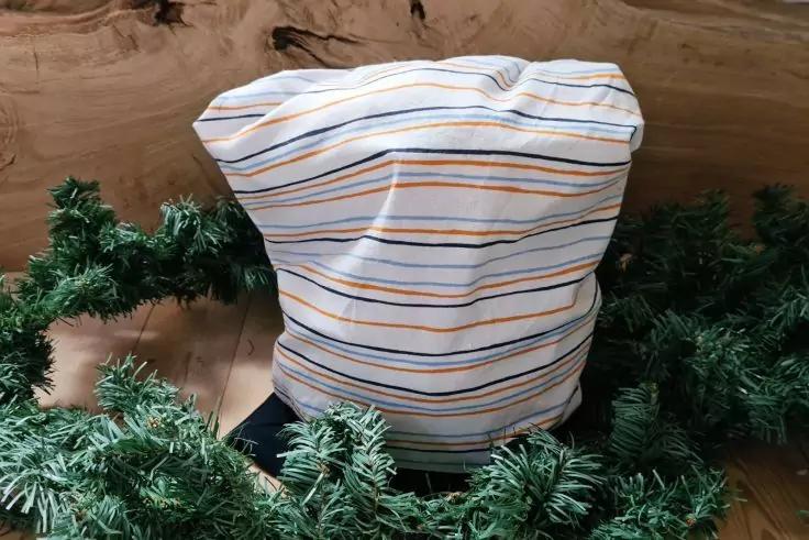 Wei&szlig;er Stoff mit horizontalen blauen, orange und grauen Streifen, zu einem aufrechten B&uuml;ndel geformt, umgeben von gr&uuml;ner k&uuml;nstlicher Tannengirlande auf hellem Holzuntergrund