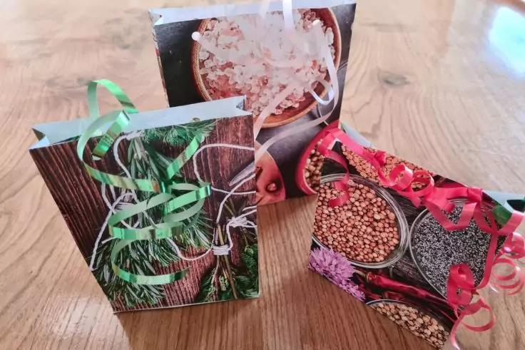 Drei kleine rechteckige Papiert&uuml;ten mit Kordelgriffen, bedruckt mit Holz-, Kr&auml;uter- und Gew&uuml;rzmotiven, mit gekringelten bunten B&auml;ndern auf einem hellen Holztisch