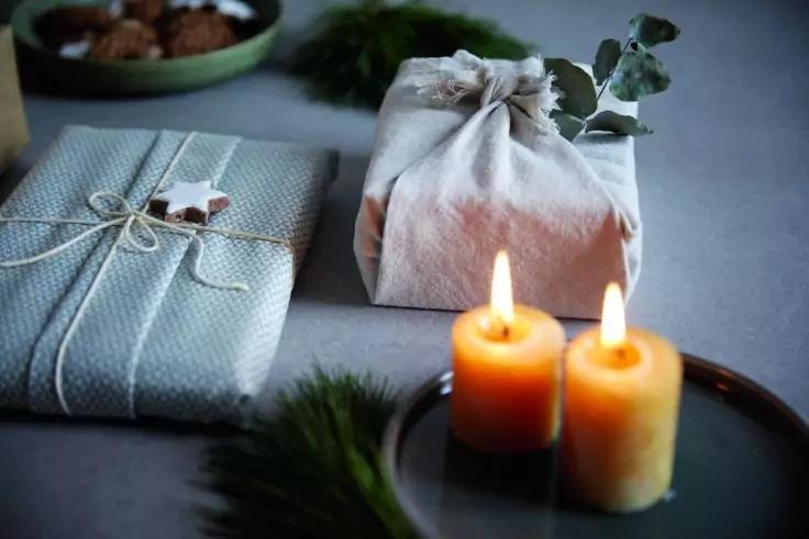 Zwei brennende Kerzen auf einem dunklen Teller, im Vordergrund zwei verpackte Geschenke mit Tannenzweigen und Eukalyptus, im Hintergrund unscharfe Schale mit Pralinen