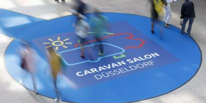 Blauer runder Bodenaufkleber mit Illustration eines Wohnwagens und einer Sonne sowie dem Text 'CARAVAN SALON D&Uuml;SSELDORF'; mehrere verschwommene Personen gehen dar&uuml;ber