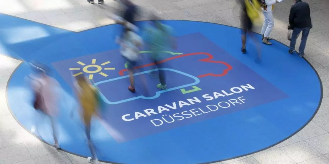 Blauer runder Bodenaufkleber mit Illustration eines Wohnwagens und einer Sonne sowie dem Text 'CARAVAN SALON D&Uuml;SSELDORF'; mehrere verschwommene Personen gehen dar&uuml;ber