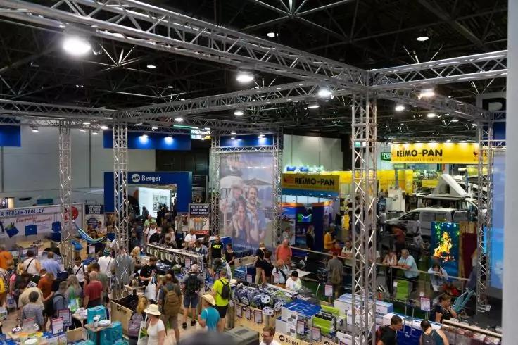 Innenaufnahme einer Messehalle mit zahlreichen St&auml;nden, Menschen, Tischen mit Waren, sichtbaren Metalltr&auml;gern an der Decke und Deckenbeleuchtung