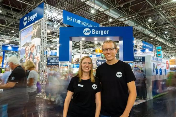 Zwei Personen in schwarzen T‑Shirts mit rundem Logo stehen vor einem blauen Messestand mit dem Schriftzug "Berger"; Messehalle mit Besuchern und weiteren St&auml;nden im Hintergrund