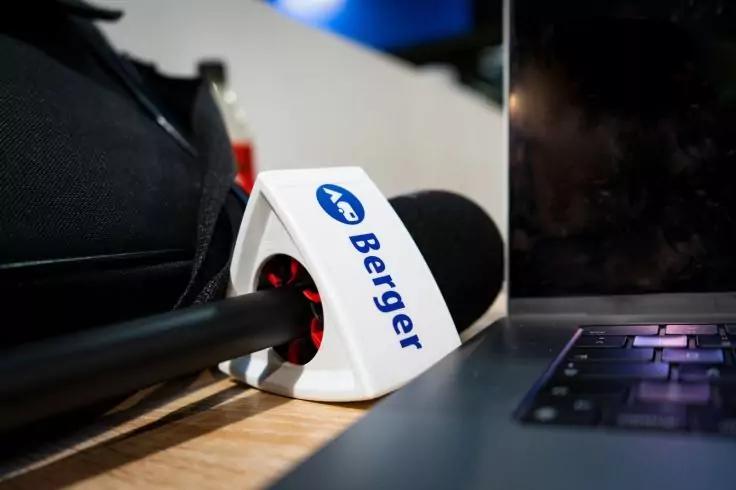 Handmikrofon mit schwarzer Schaumstoffhaube und wei&szlig;em Mikrofonw&uuml;rfel mit blauem Schriftzug 'Berger' auf einer Holzoberfl&auml;che neben einer Laptop-Tastatur; im unscharfen Hintergrund eine schwarze Tasche und ein Bildschirm
