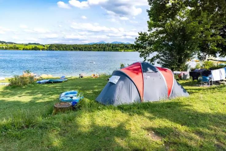 Rotes-graues Zelt auf grasigem Seeufer, zwei auf dem Gras liegende Kajaks, Baum rechts, See und bew&ouml;lkter Himmel im Hintergrund