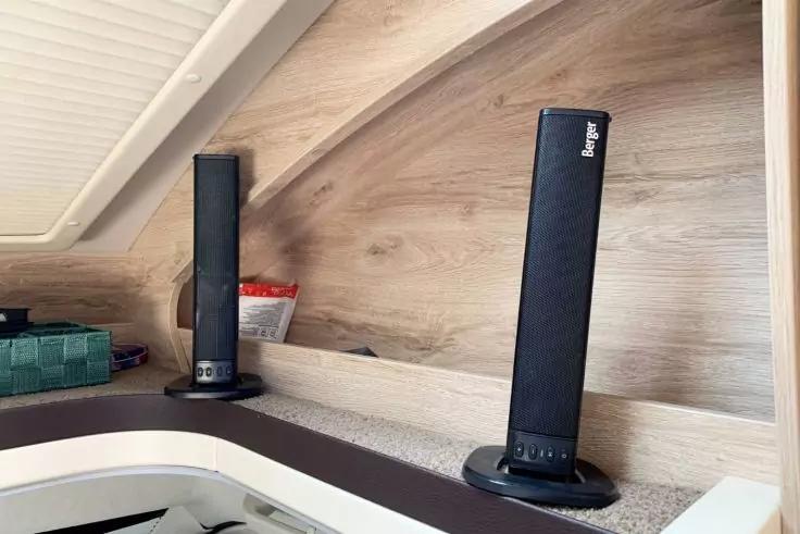 Zwei schwarze, vertikale Lautsprecher auf runden Sockeln auf einem hellen Regal vor einer schr&auml;gen Holzverkleidung; links ein gr&uuml;nes K&auml;stchen und ein kleines rot-wei&szlig;es T&uuml;tchen