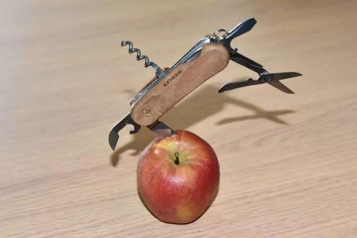 Klappmesser mit mehreren ge&ouml;ffneten Werkzeugen (Korkenzieher, Schere) auf einem roten Apfel auf einer hellen Holzfl&auml;che