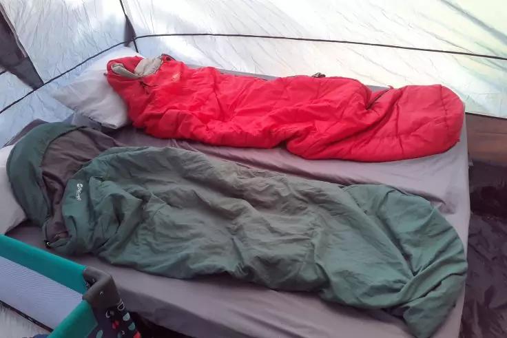 Zwei Schlafs&auml;cke (rot oben, gr&uuml;n unten) auf einer grauen Matratze im Zelt; unten links ein t&uuml;rkisfarbenes Kunststoffteil sichtbar