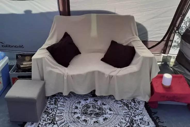 Beige &uuml;berzogene zweisitzige Couch mit zwei dunklen Kissen, davor schwarz-wei&szlig;es Mandala-Teppichmuster, links grauer Hocker, rechts roter Beistelltisch mit wei&szlig;em zylindrischem Gegenstand, heller Stoffhintergrund mit dunklen Stangen