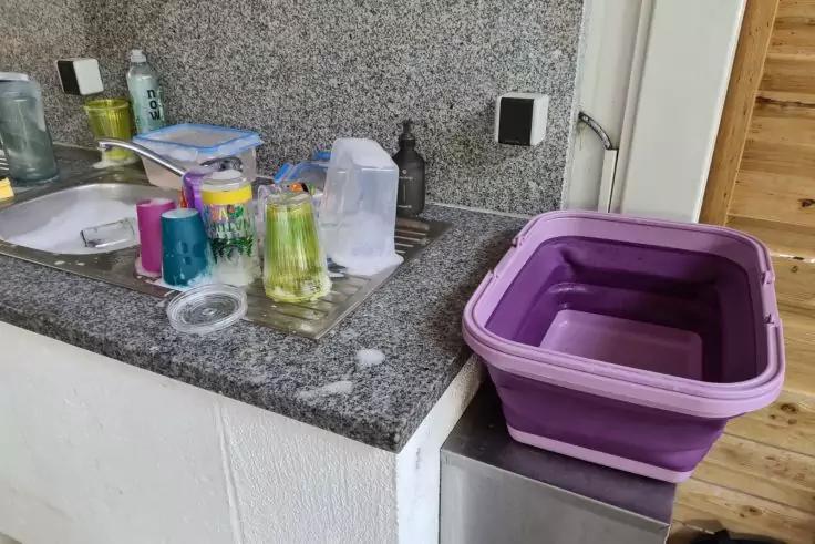 K&uuml;chenarbeitsplatte mit Sp&uuml;le und Wasserhahn, Becher, Flaschen, Schwamm und Lappen auf der Arbeitsplatte, zusammengefaltete lila faltbare Plastikwanne auf einem Edelstahlger&auml;t, Steckdose an der Wand