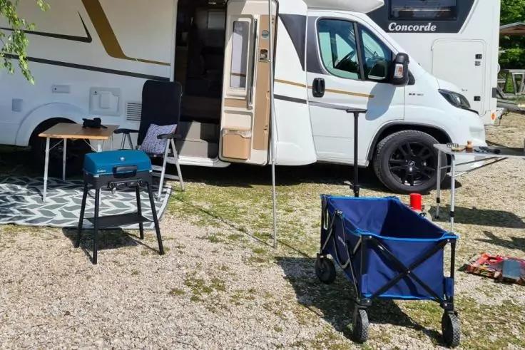 Wei&szlig;es Wohnmobil mit ge&ouml;ffneter T&uuml;r und ausgefahrenem Sonnendach; davor kleiner Tisch mit tragbarem Grill, Klappstuhl und blauer faltbarer Bollerwagen auf Kies