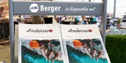 Prospektst&auml;nder mit Schild "Berger in Kooperation mit" und zwei Magazinexemplaren mit Titel "Anderswo", auf den Titeln zwei Personen neben t&uuml;rkisfarbenem Wasser; im Hintergrund Regale eines Gesch&auml;fts