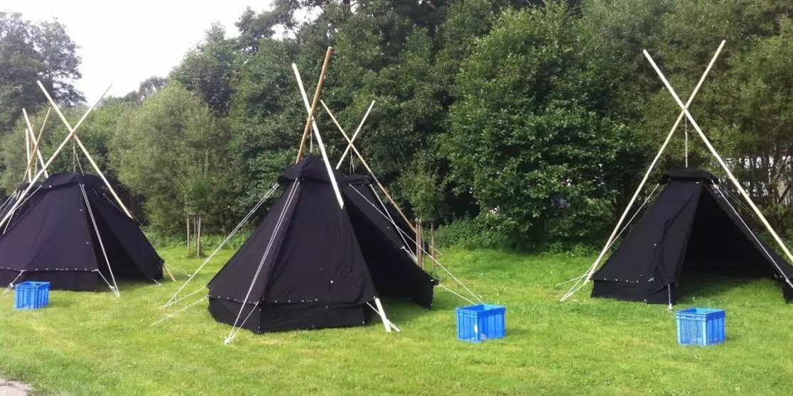 Drei schwarze Tipi-Zelte mit gekreuzten Holzstangen auf einer Wiese, davor blaue Kunststoffkisten, im Hintergrund B&auml;ume