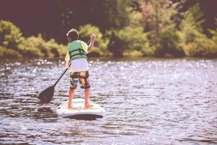 Person steht auf einem Stand-up-Paddleboard mit Schwimmweste und Paddel, auf dem Wasser, Ufer mit B&auml;umen im Hintergrund