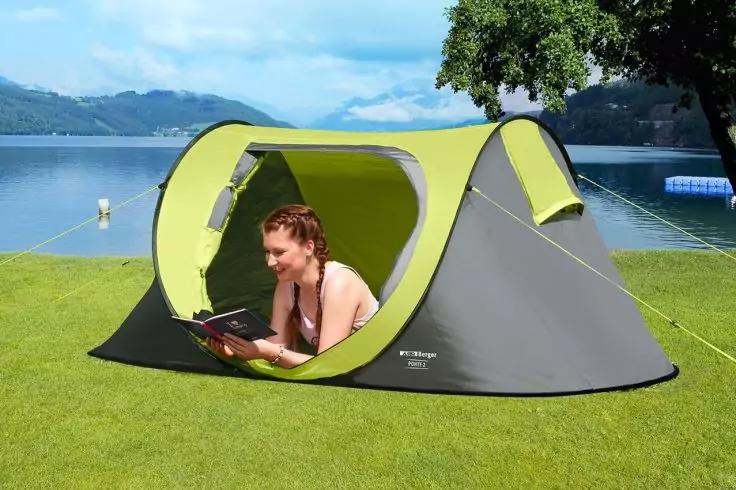 Person liegt in einem kleinen graugr&uuml;nen Pop-up-Zelt auf Gras und liest ein Buch; im Hintergrund See, Berge und ein Baum