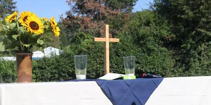 Tisch mit wei&szlig;er Tischdecke und dunkelblauem L&auml;ufer, h&ouml;lzernes Kreuz in der Mitte, zwei Gl&auml;ser, Sonnenblumen in einer Vase, gr&uuml;ne Hecke im Hintergrund