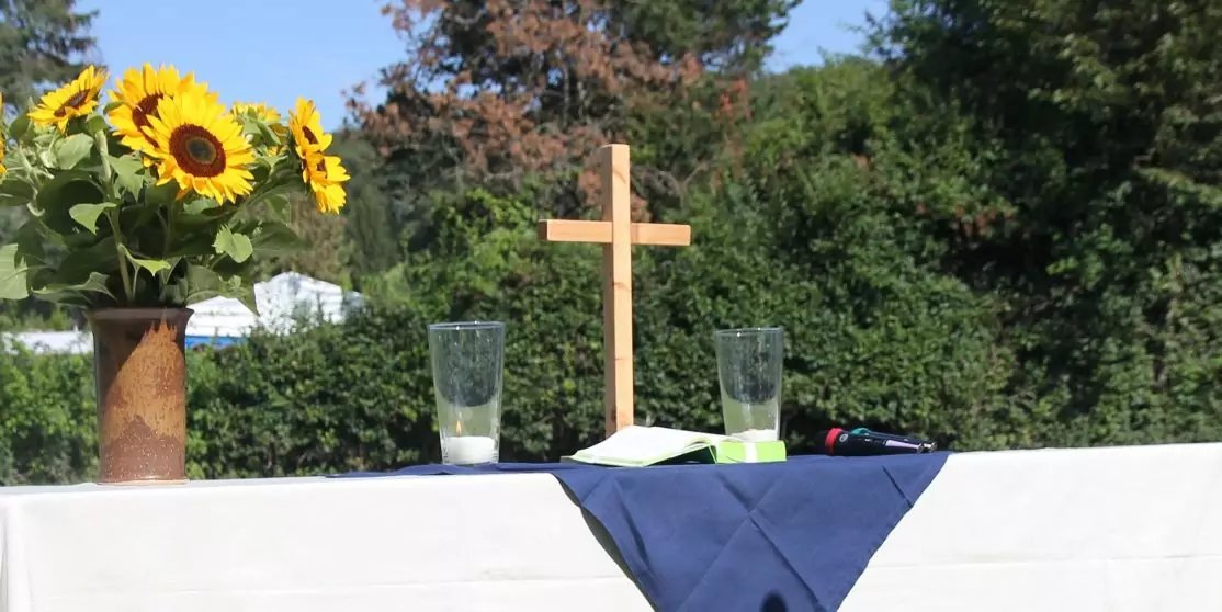 Tisch mit wei&szlig;er Tischdecke und dunkelblauem L&auml;ufer, h&ouml;lzernes Kreuz in der Mitte, zwei Gl&auml;ser, Sonnenblumen in einer Vase, gr&uuml;ne Hecke im Hintergrund