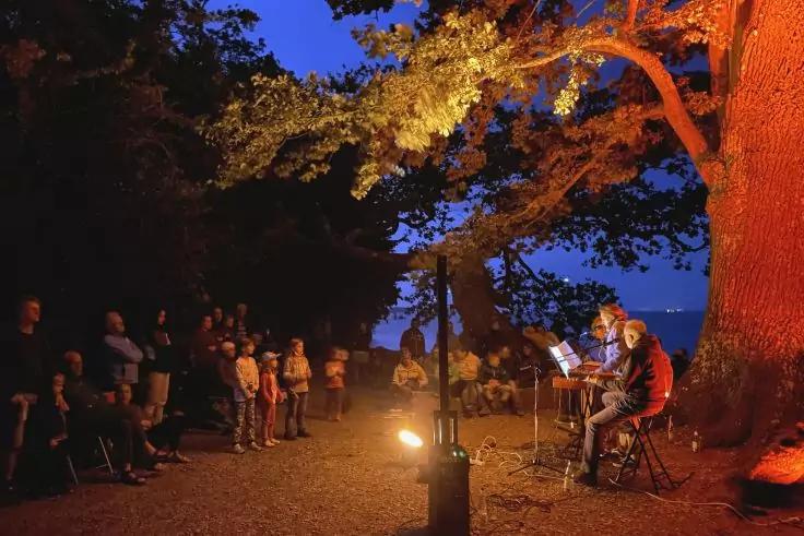 Menschen stehen links; rechts zwei sitzende Musiker mit Gitarren und Notenst&auml;ndern unter einem gro&szlig;en Baum, der orange beleuchtet ist; blauer Abendhimmel und Wasser im Hintergrund; kleines Bodenlicht
