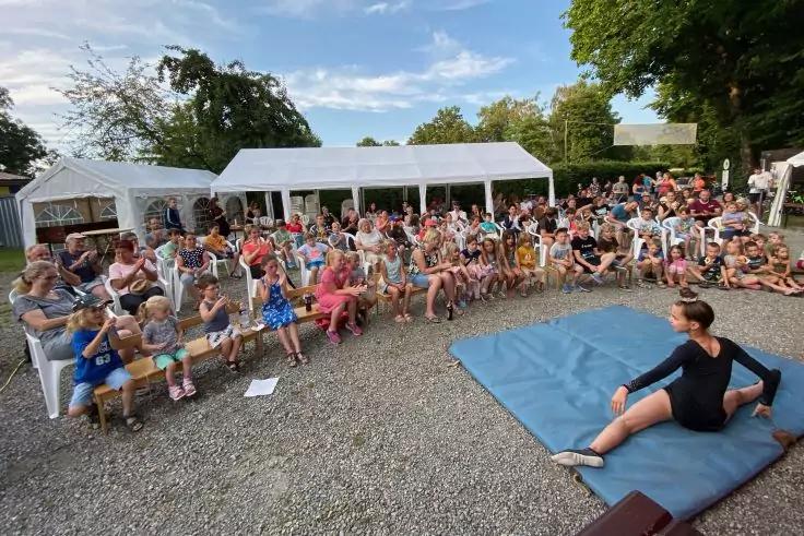 Viele Menschen sitzen auf B&auml;nken vor wei&szlig;en Pavillons; eine Person in schwarzer Kleidung sitzt auf einer blauen Matte; Kiesboden, B&auml;ume und blauer Himmel sichtbar