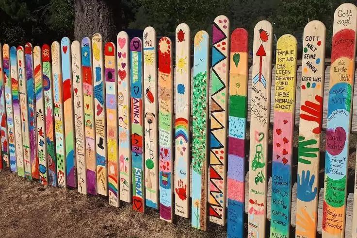 Bunte, einzeln bemalte Holzlatten eines Lattenzauns, im Hintergrund B&auml;ume