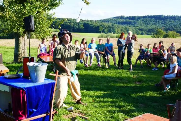 Person in gr&uuml;n-brauner Kleidung und Hut steht auf einer Wiese neben Tischen, Eimern und Requisiten. Eine Gruppe von Menschen sitzt und steht im Halbkreis und blickt zur Person. Im Hintergrund B&auml;ume, Feld und H&uuml;gel unter blauem Himmel