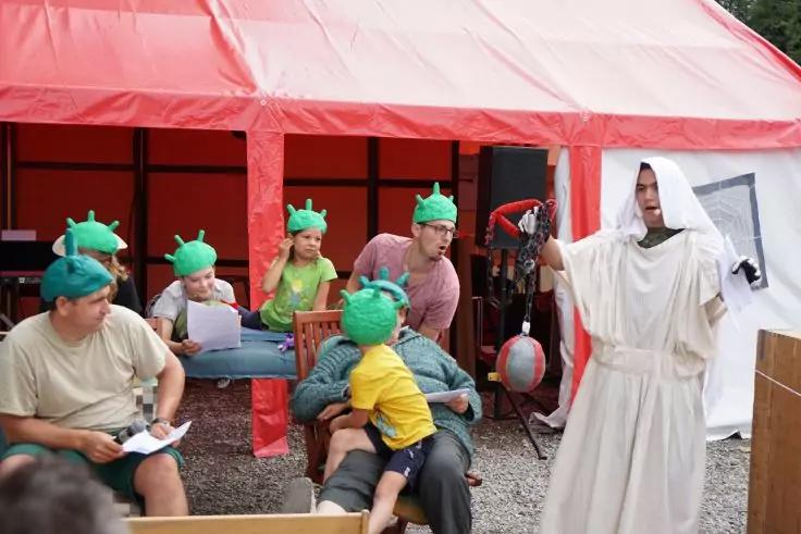 Mehrere Personen sitzen vor einem roten Zelt; einige tragen gr&uuml;ne Kopfbedeckungen mit Stacheln. Eine Person in wei&szlig;er Robe steht und h&auml;lt ein schwarzes Ger&auml;t mit rotem Kabel; Papiere und St&uuml;hle sind sichtbar