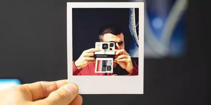 Hand h&auml;lt ein Polaroid-Foto; im Foto eine Person, die eine Sofortbildkamera vor das Gesicht h&auml;lt, dunkler Hintergrund