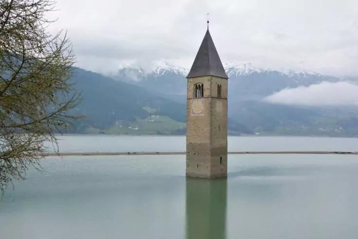 Steinturm mit spitzem Dach steht teilweise im ruhigen See, Spiegelung im Wasser, im Hintergrund schneebedeckte Berge und bew&ouml;lkter Himmel, links ein Baumzweig