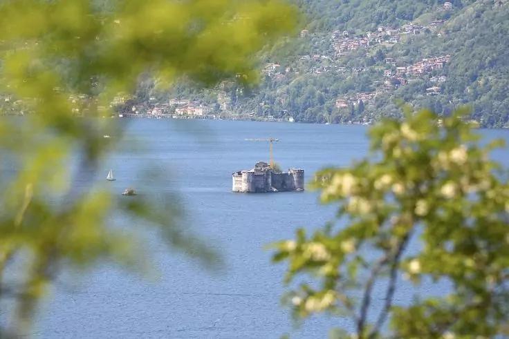 Kleines steinernes Geb&auml;ude mit Fahnenmast auf einer kleinen Insel in einem See, unscharfe Zweige im Vordergrund, bewaldeter Hang mit H&auml;usern im Hintergrund
