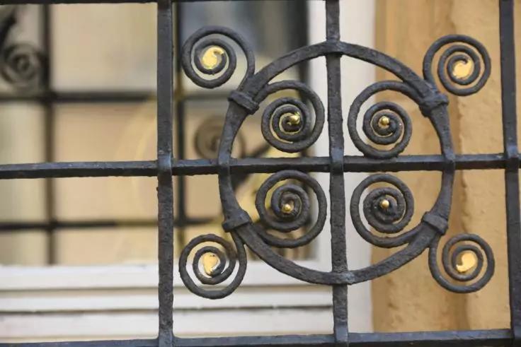 Nahaufnahme eines metallenen Gitters mit kreisf&ouml;rmigen Spiralmotiven vor einem Fenster und einer beigen Wand