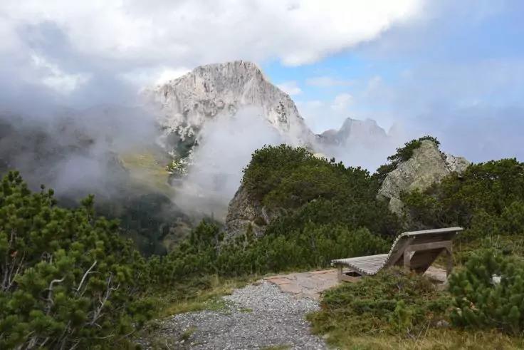 Bank neben einem Schotterweg, im Vordergrund Str&auml;ucher, felsige Berggipfel teilweise von Nebel und Wolken bedeckt, blauer Himmel mit Wolken
