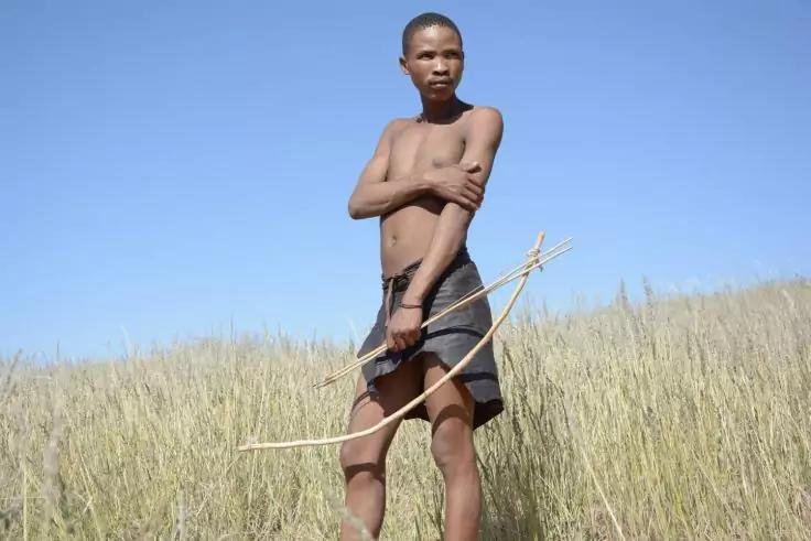 Person steht in hohem Gras, oberk&ouml;rperfrei, Lendentuch, h&auml;lt einen Holzbogen und Pfeile, blauer Himmel