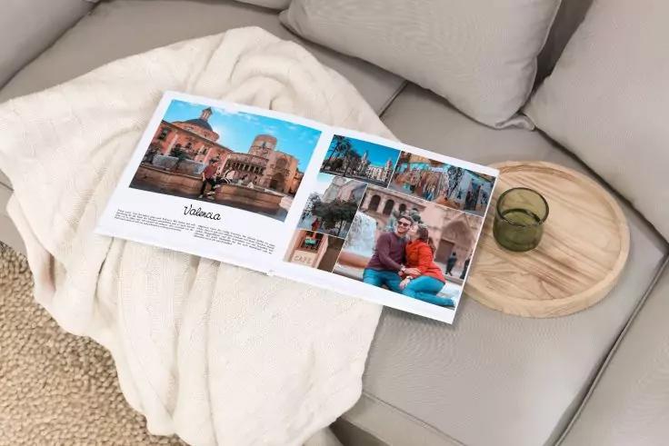 Aufgeschlagenes Fotobuch mit Bildern von Geb&auml;uden und einer Person auf einem hellen Sofa mit heller Decke, daneben ein rundes Holztablett mit gr&uuml;nem Glas